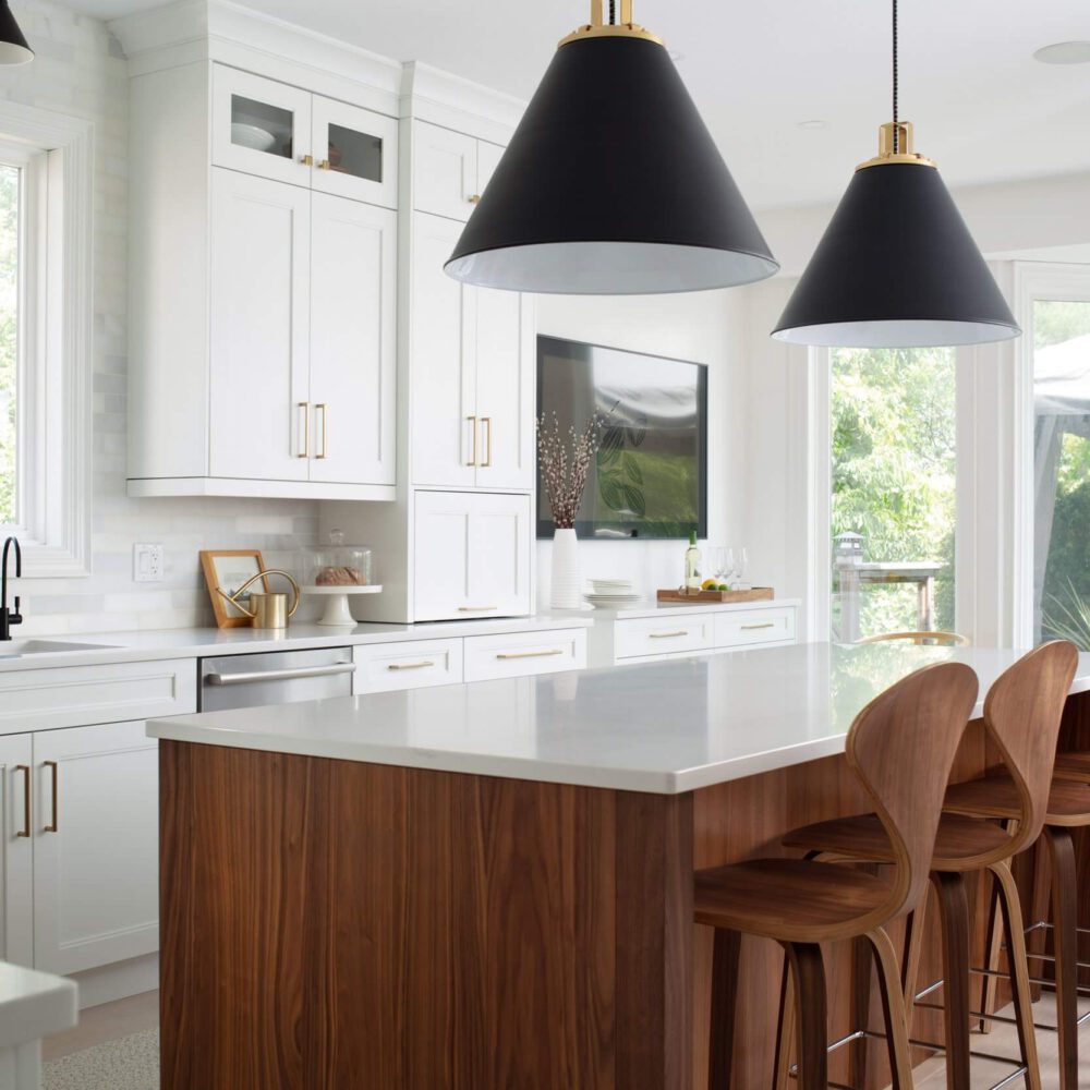 Bright white kitchen with walnut island, black pendant lights, brass accents, and tall stools beside a sunlit window.