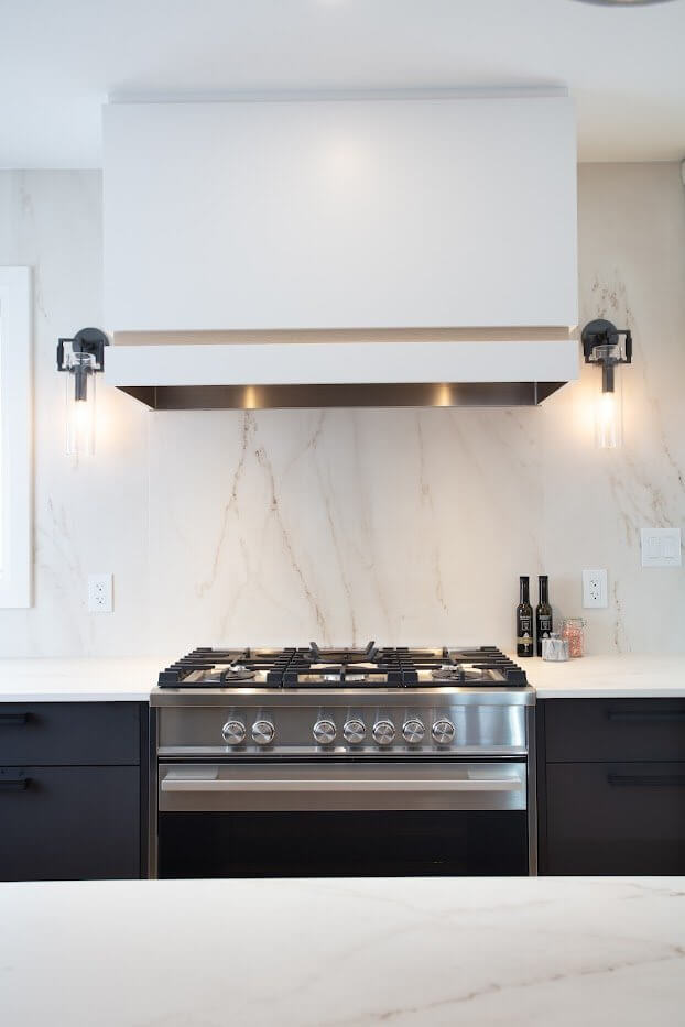 Modern kitchen with a stainless gas range, white marble-look backsplash, black cabinets, and matching wall sconces.