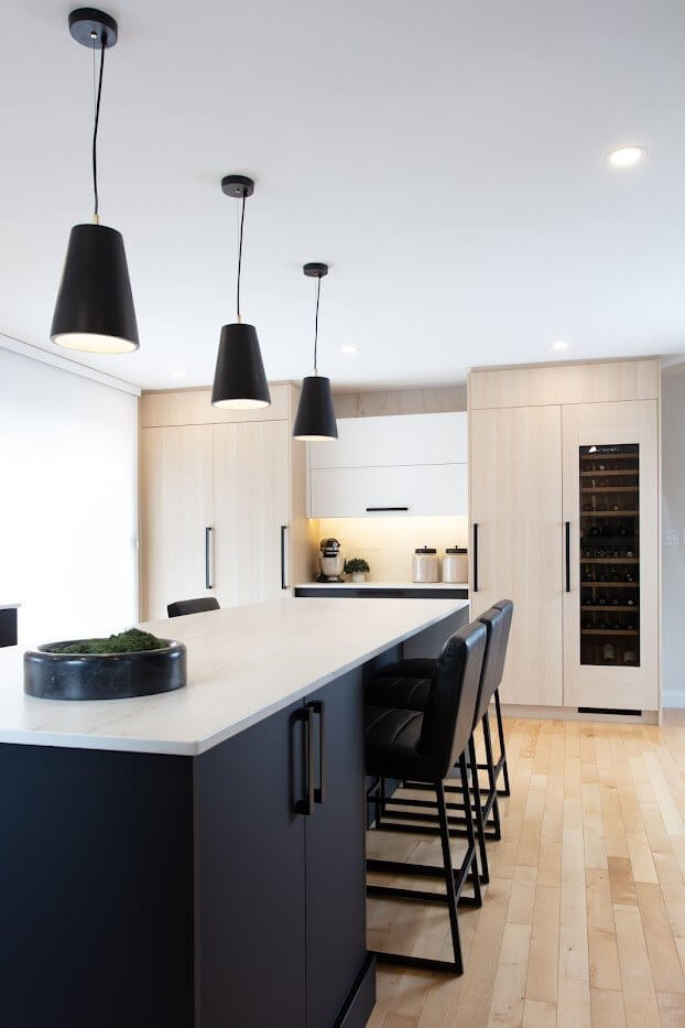 Modern kitchen with pale wood cabinetry, a white island, black pendant lights, and a built-in wine fridge.