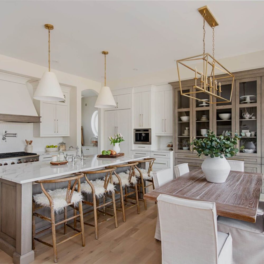 Byron Beauty kitchen by Casey’s Kitchens featuring muted white cabinetry, gray-stained oak accents, a marble-like island, brass pendant lighting, and a glass-front pantry cabinet