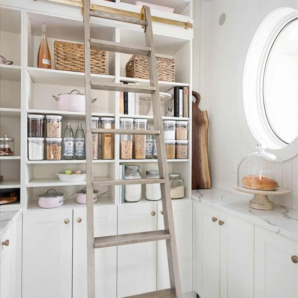 Byron Beauty kitchen pantry by Casey's Kitchens featuring white cabinetry, a freestanding wooden ladder, glass jars with dry goods, woven baskets, and a round window.