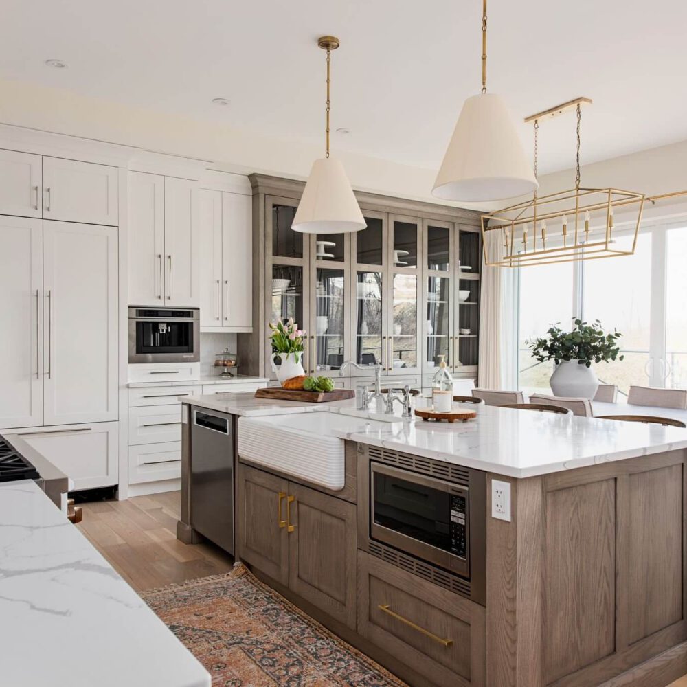 Modern white and light wood kitchen with a large central island, glass-front display cabinets, stainless steel appliances, pendant lights, and sliding glass doors