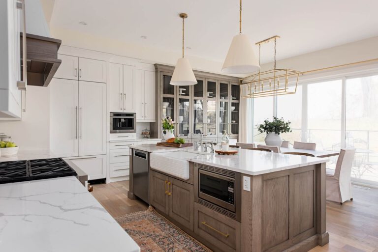 Modern white and light wood kitchen with a large central island, glass-front display cabinets, stainless steel appliances, pendant lights, and sliding glass doors