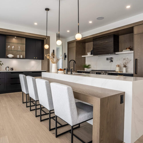 Modern kitchen with ebony cabinetry, warm oak framing, a quartz island, and three pendant lights over upholstered stools.