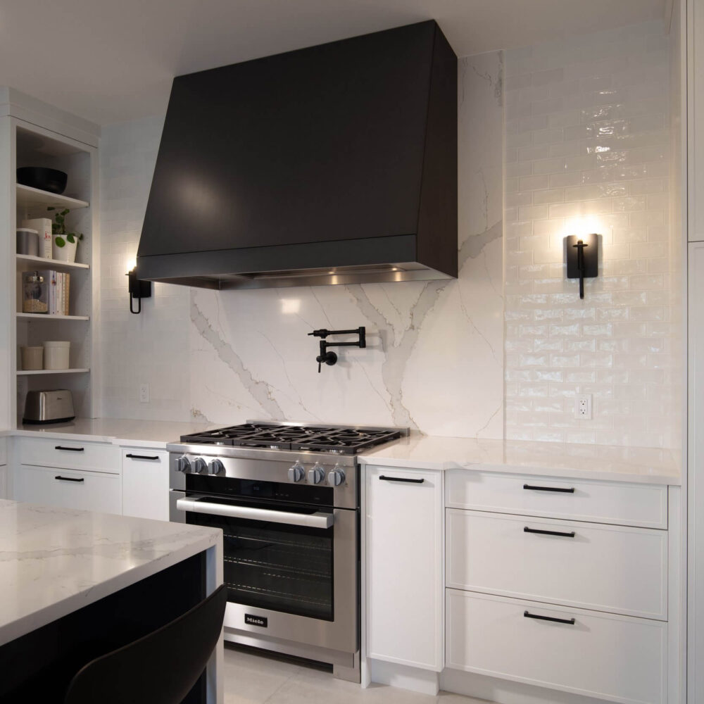 Modern white kitchen with a bold black range hood, marble-look backsplash, stainless stove and black hardware.