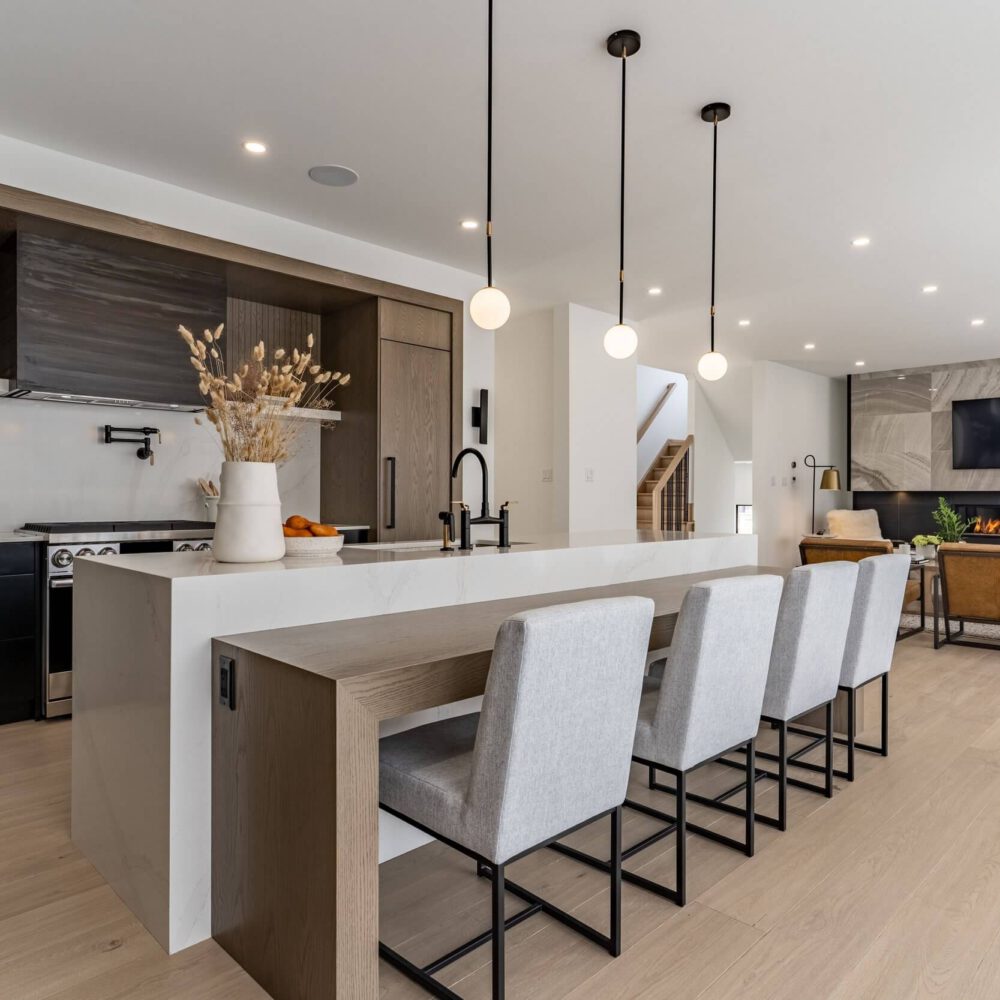 A modern kitchen with dark cabinetry, a large quartz island, pendant lights and a matching living room fireplace wall.