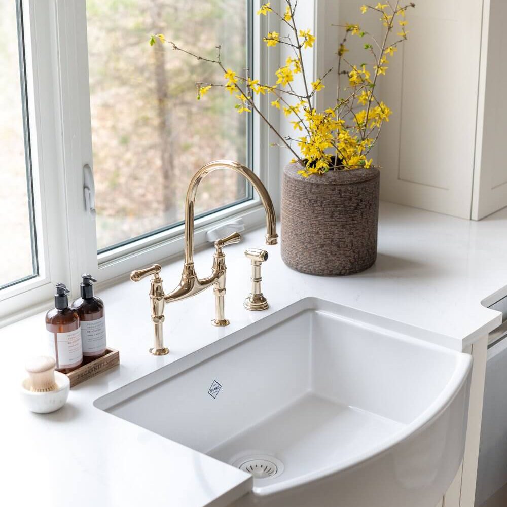 White farmhouse sink with a gold faucet and a vase of yellow flowers on a countertop.