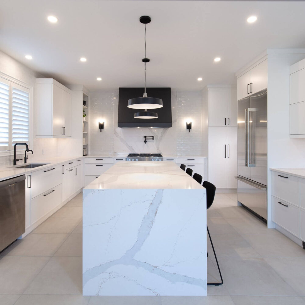 Bright white kitchen with a large marble island, black pendant lights, built-in appliances, and a sleek black range hood.