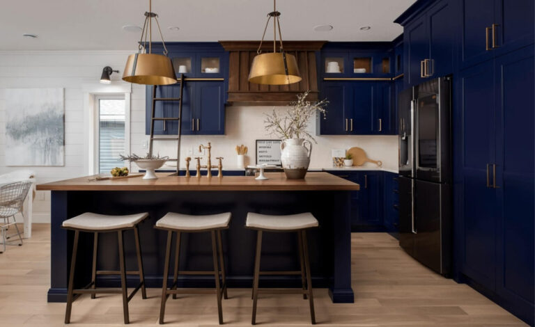 Modern kitchen with navy blue cabinetry, wooden accents, and a central island with three stools. Brass light fixtures add elegance.