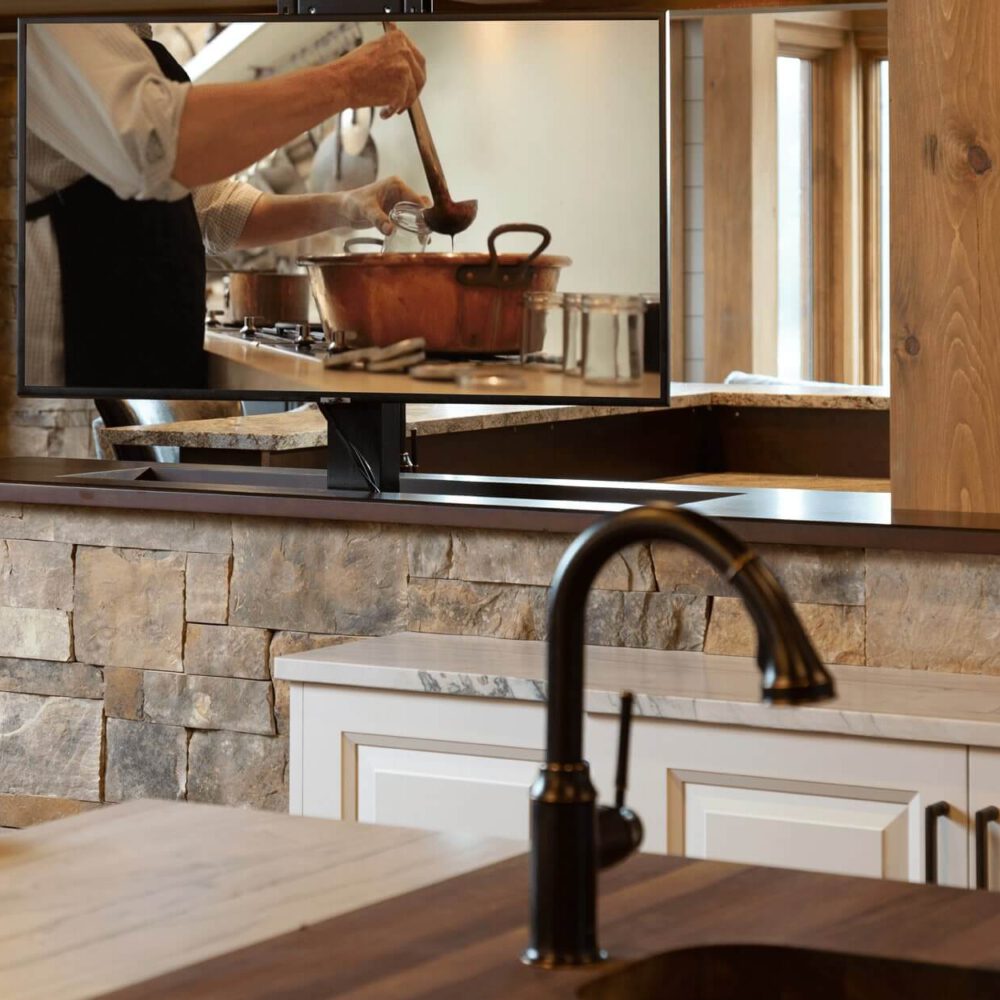 Kitchen interior with a television displaying a person stirring a pot, featuring wooden cabinetry and a stone countertop.