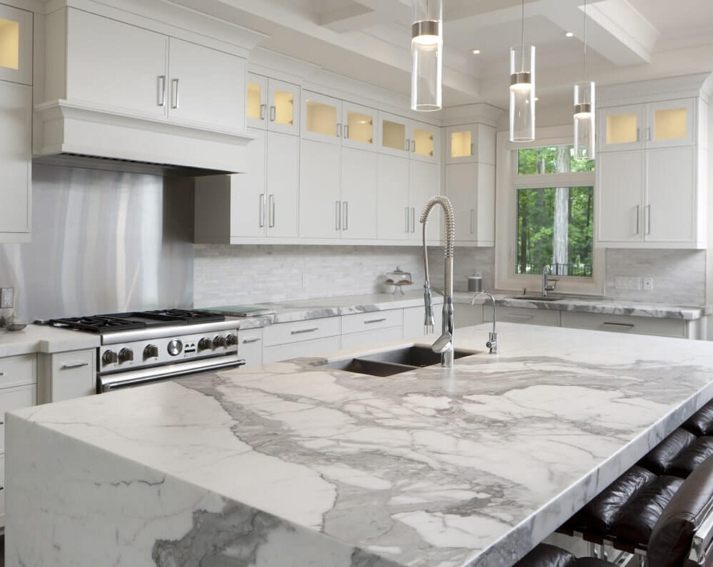 Modern kitchen with white cabinetry, a large marble island, and stainless steel appliances, featuring pendant lighting and a sleek design.