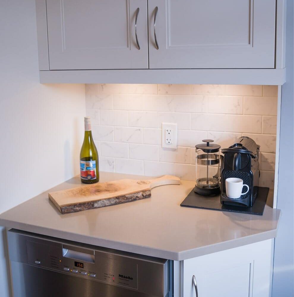 Old South Kitchen area with white shaker cabinets, walnut floating shelves, under-cabinet lighting, a small wine bottle, coffee station, and a Miele dishwasher beneath a light countertop.