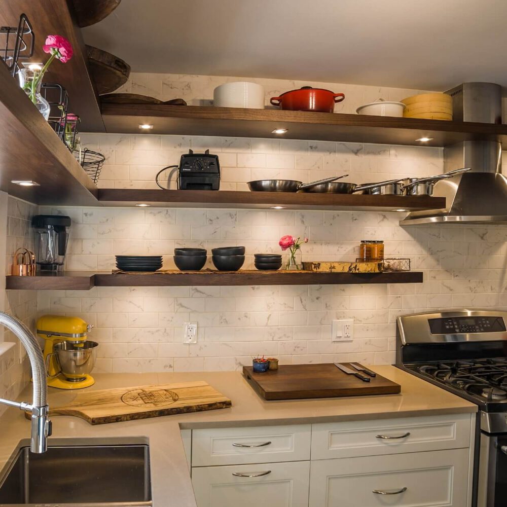 Old South Kitchen by Casey’s Kitchens featuring walnut floating shelves, white tile backsplash, a bright yellow stand mixer, and a stainless steel gas range with a cutting board on the counter.