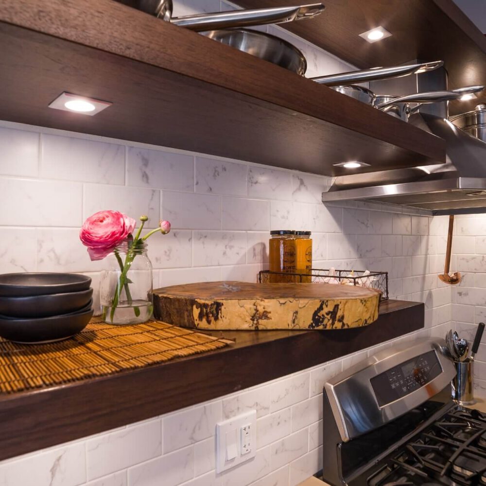OldSouth kitchen scene with white marble tile backsplash, dark wood open shelves, a stainless range hood, a round wooden cutting board, and a glass vase with pink ranunculus alongside spice jars.