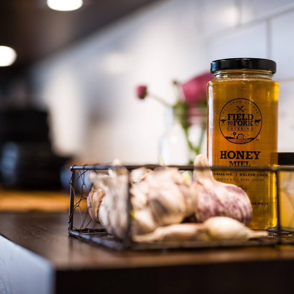 Old South Kitchen scene with a tall golden honey jar labeled Field to Fork and a smaller Field to Fork mustard jar on a dark wood counter, garlic bulbs in a wire basket, white subway tile backsplash