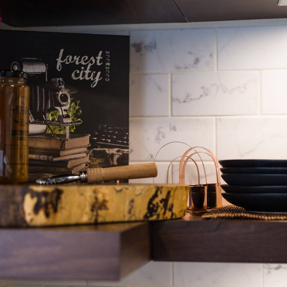 Old South Kitchen display with a dark walnut shelf, copper utensil accents, stacked black plates, glass bottle and a chalkboard-style forest city cookbook against a white marble tile backsplash