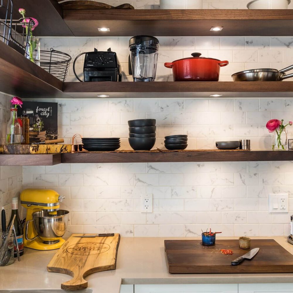 Old South Kitchen by Casey's Kitchens features walnut floating shelves above a white marble subway tile backsplash, with a yellow mixer on the counter and black dishware stacked on shelves.