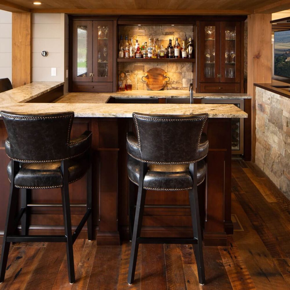 Rustic wooden bar cabinetry at Fruit Ridge with stone feature wall, glass-front storage, liquor bottles, and dark leather bar stools.
