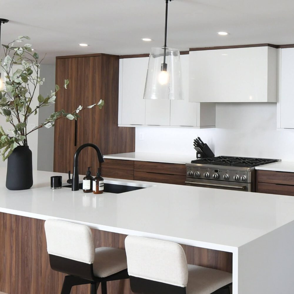 Modern contemporary kitchen by Casey's Kitchens with white flat-panel cabinets, dark wood lower cabinetry, a large white quartz island, a black faucet, and a tall plant centerpiece.