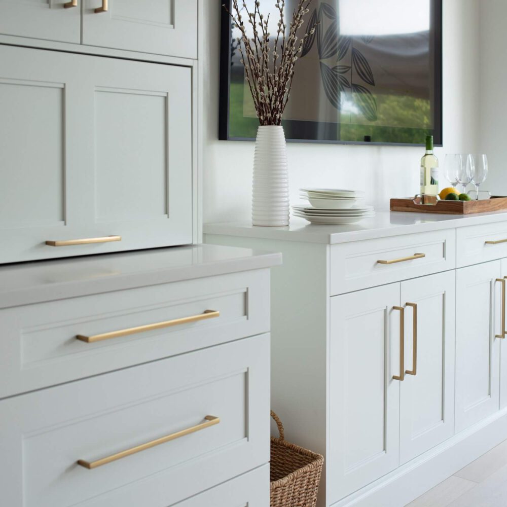 White shaker kitchen cabinetry with brass handles, a walnut-toned cabinet, and a bright window beside styled decor.