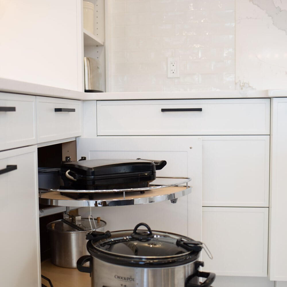 Pull-out corner pantry shelves hold a Crock-Pot slow cooker and cookware inside a white kitchen with glossy tile backsplash.