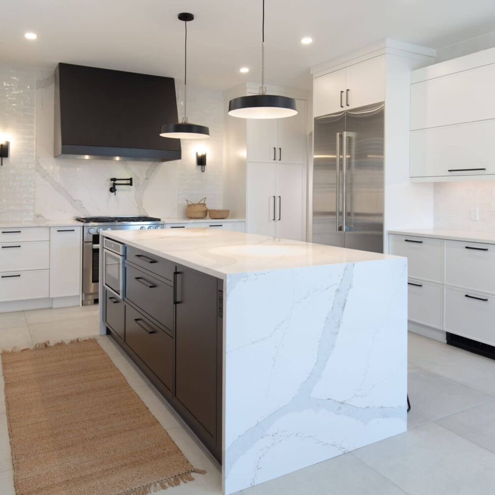 Modern white kitchen with a large marble island, black range hood, stainless steel fridge, and brass woven rug.