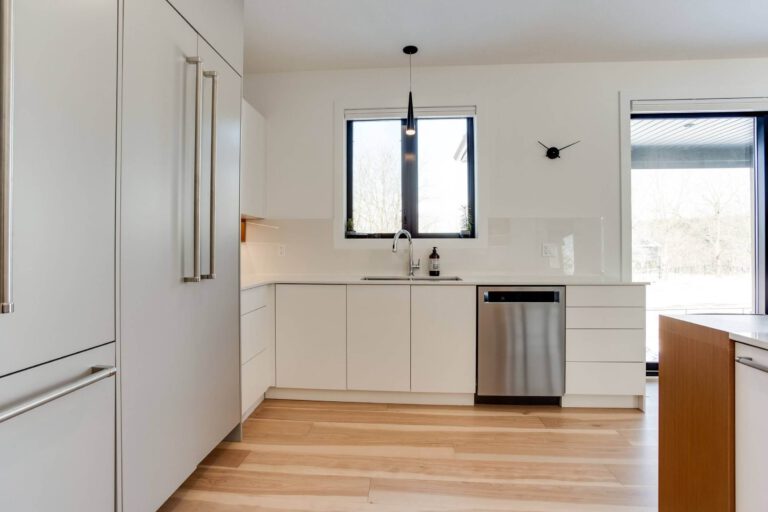 Modern kitchen with white cabinetry, stainless steel dishwasher, and large windows providing natural light.