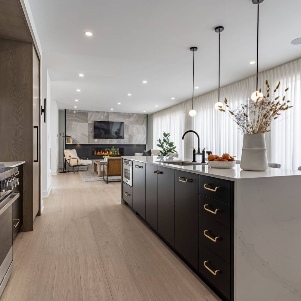 Modern kitchen with dark cabinetry, a white waterfall island, pendant lights and a living area with fireplace beyond.