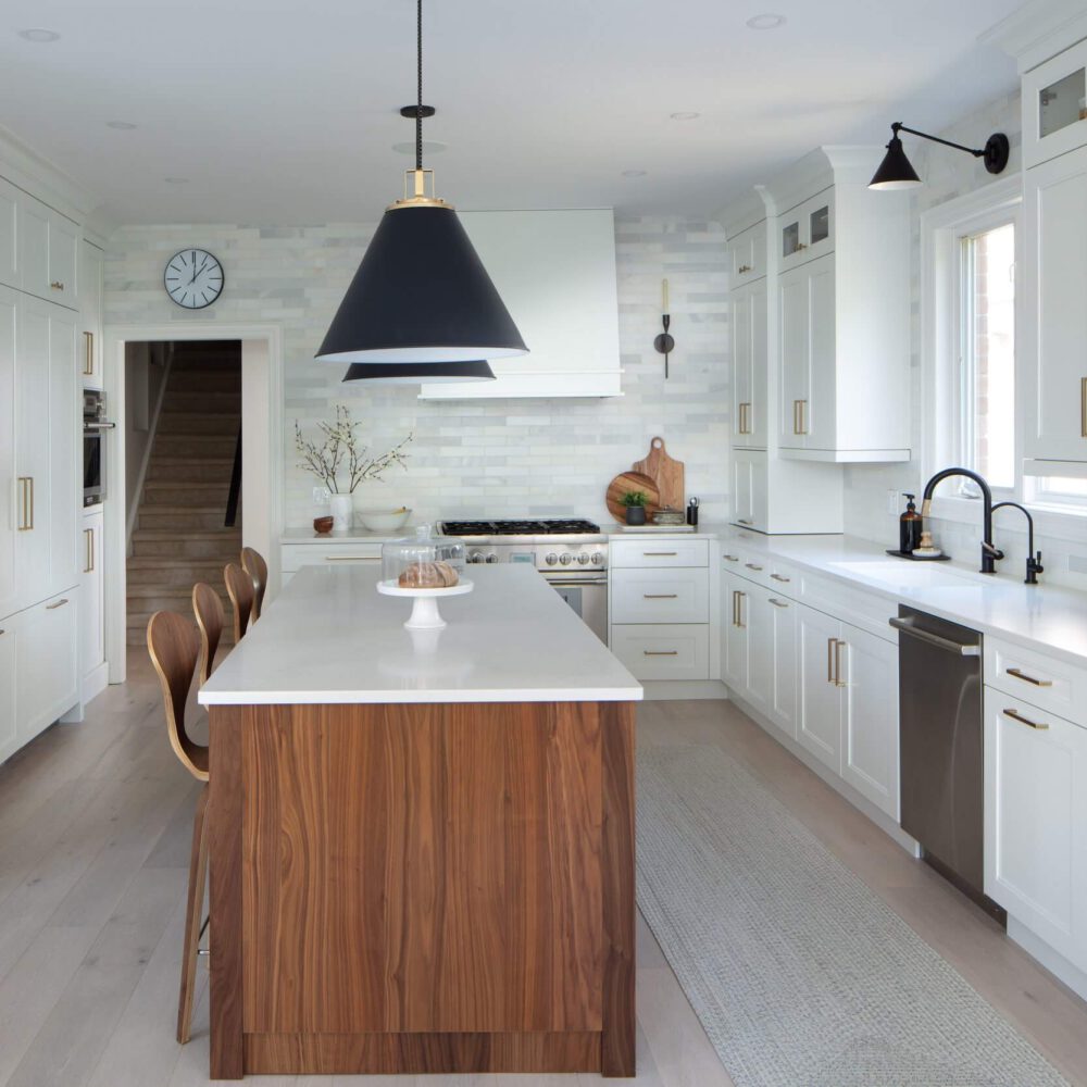 Bright white kitchen with a walnut island, black pendant lights, gold hardware, and a long counter beside tall cabinets.