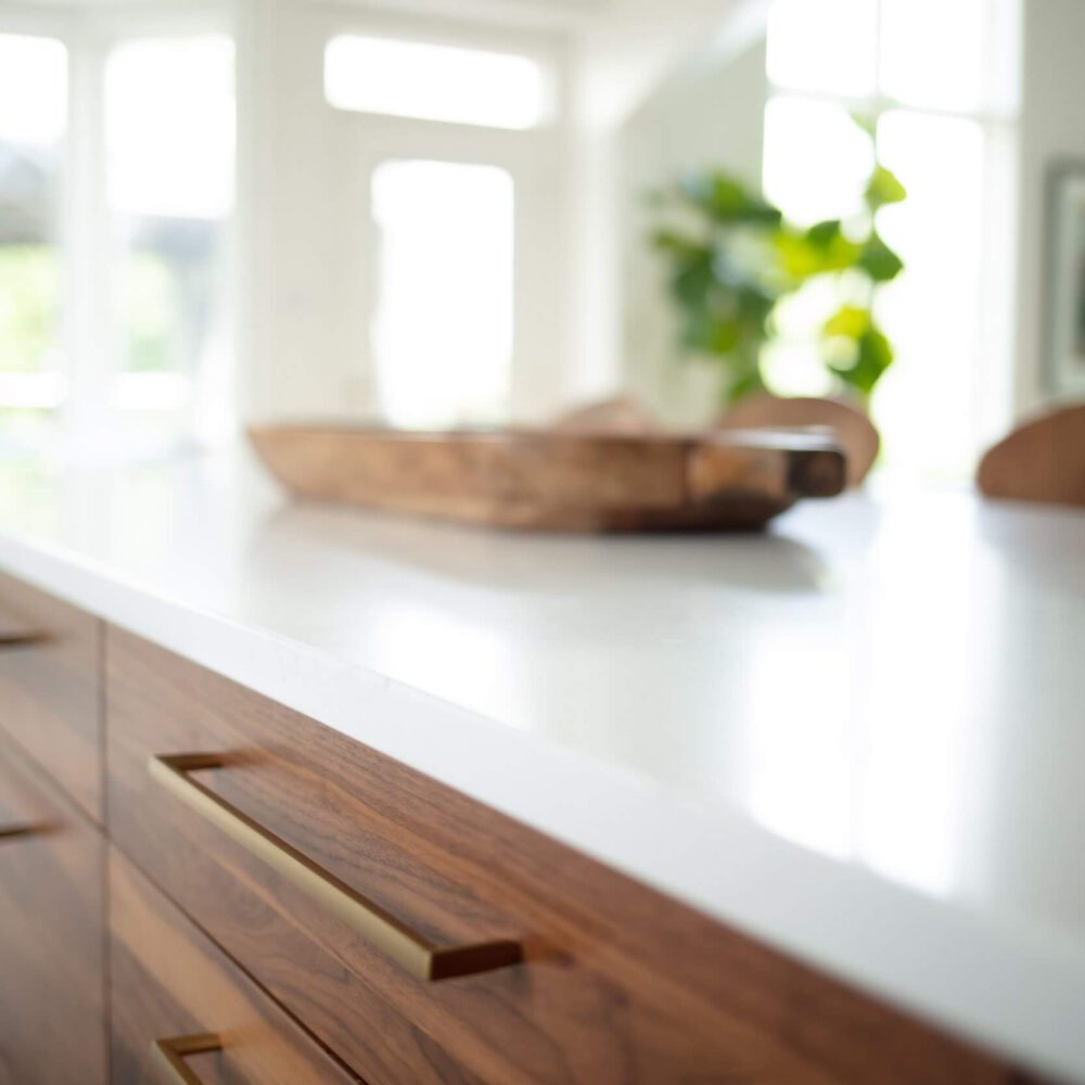 Walnut cabinetry and a white countertop sit beside bright windows, with a wooden bowl and blurred greenery behind.