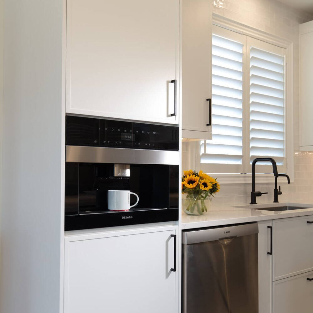 Tall white kitchen cabinetry frames a built-in Miele oven beside a sink, with black fixtures and shuttered window light.