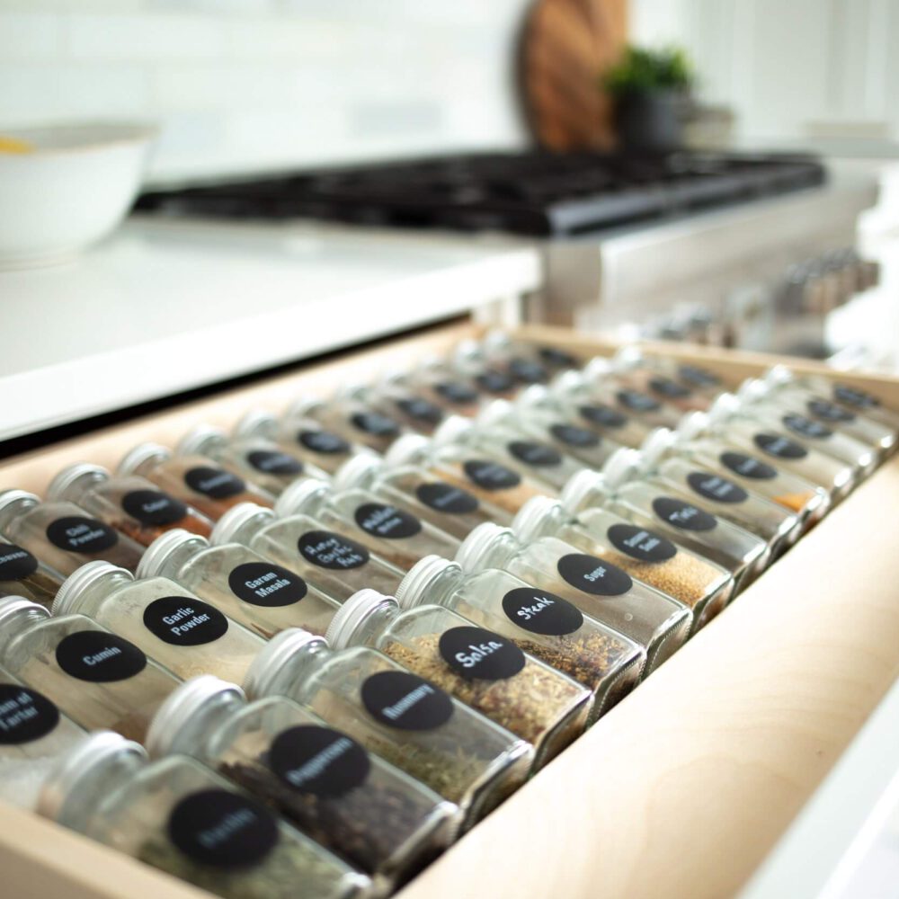 Open kitchen drawer filled with labeled spice jars beside a white countertop and a stove in a bright modern kitchen.