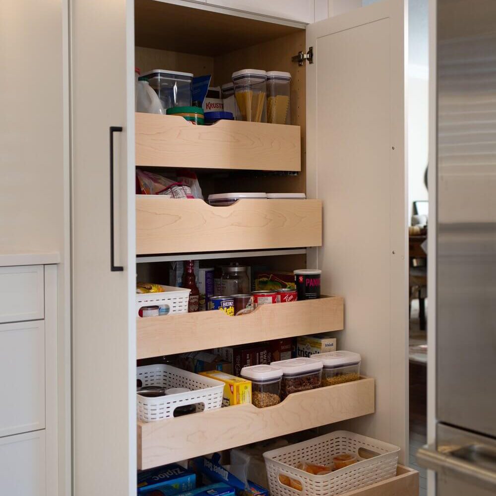 Tall kitchen pantry cabinet with pull-out drawers, stocked with dry goods, jars and baskets beside a stainless steel fridge.