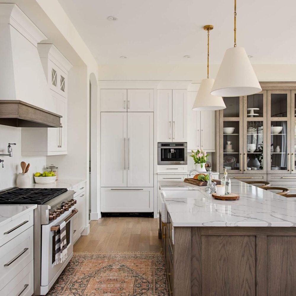Byron Beauty kitchen: white cabinets with natural wood island, marble countertops, brushed gold hardware, and a glass-front cabinet wall, in a modern farmhouse style by Casey’s Kitchens.