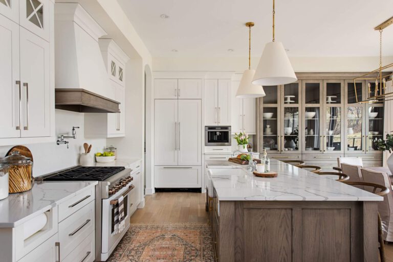 Byron Beauty kitchen: white cabinets with natural wood island, marble countertops, brushed gold hardware, and a glass-front cabinet wall, in a modern farmhouse style by Casey’s Kitchens.
