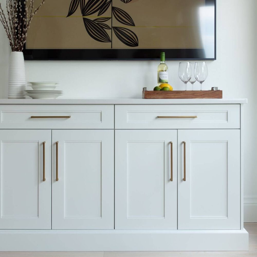 White shaker-style cabinetry with brass pulls sits beside a bright window, styled with dishes, fruit, and wine glasses.