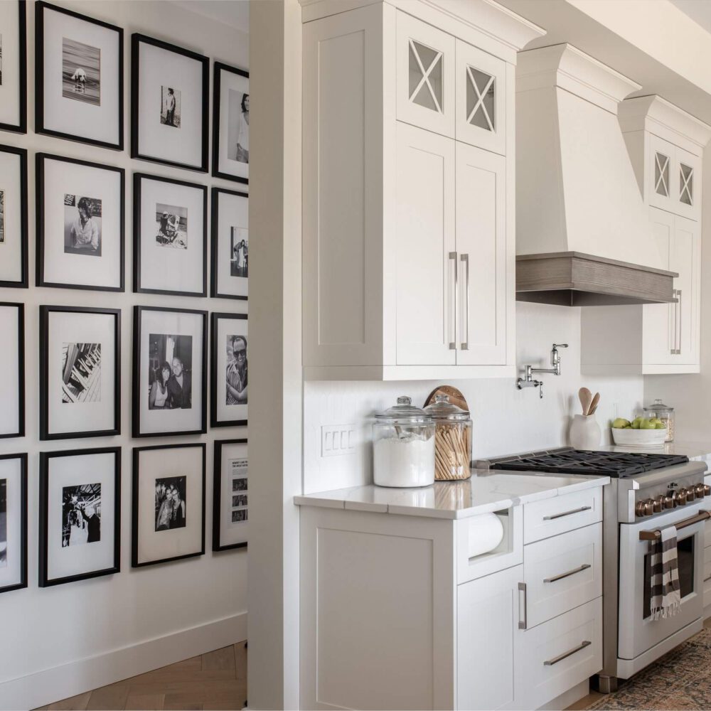 White shaker kitchen with a wall of black-framed photo gallery on the left, integrated white cabinetry, stainless steel stove, and light wood floors from Casey's Kitchens' Byron Beauty project in London, Ontario