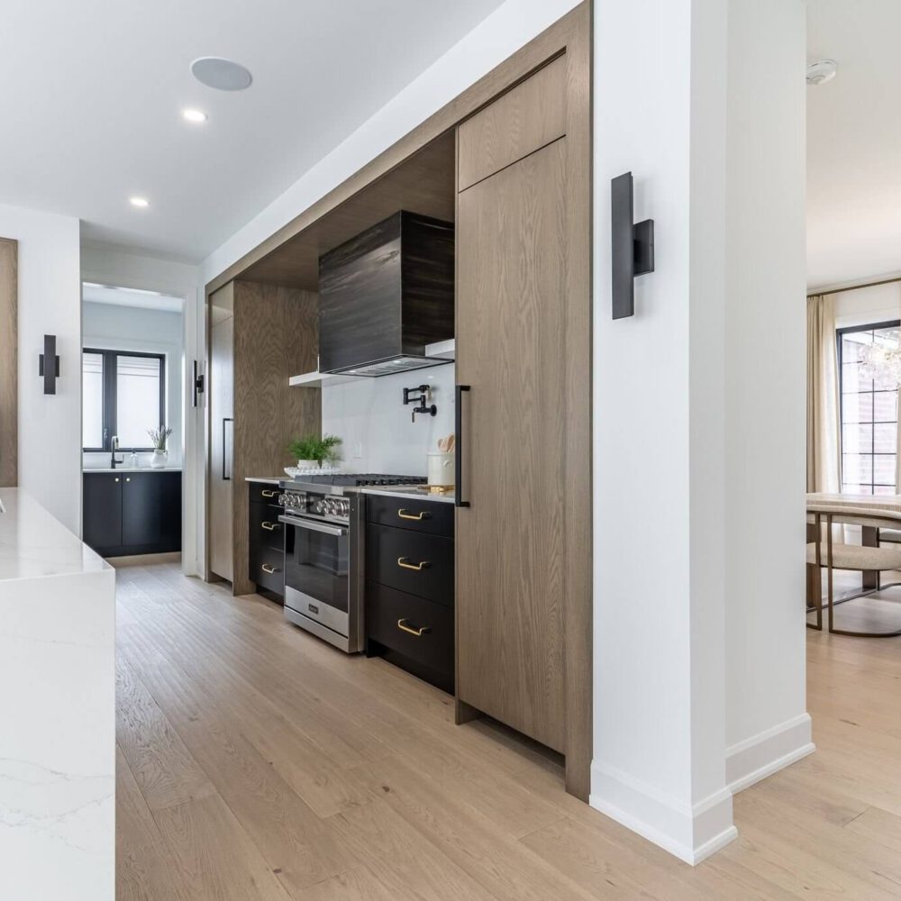 Open-plan kitchen with dark cabinetry, warm oak paneling, a white quartz island, and a dining area with a sculptural chandelier.