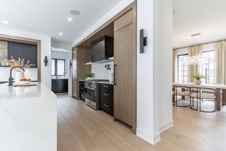 Open-plan kitchen with dark cabinetry, warm oak paneling, a white quartz island, and a dining area with a sculptural chandelier.