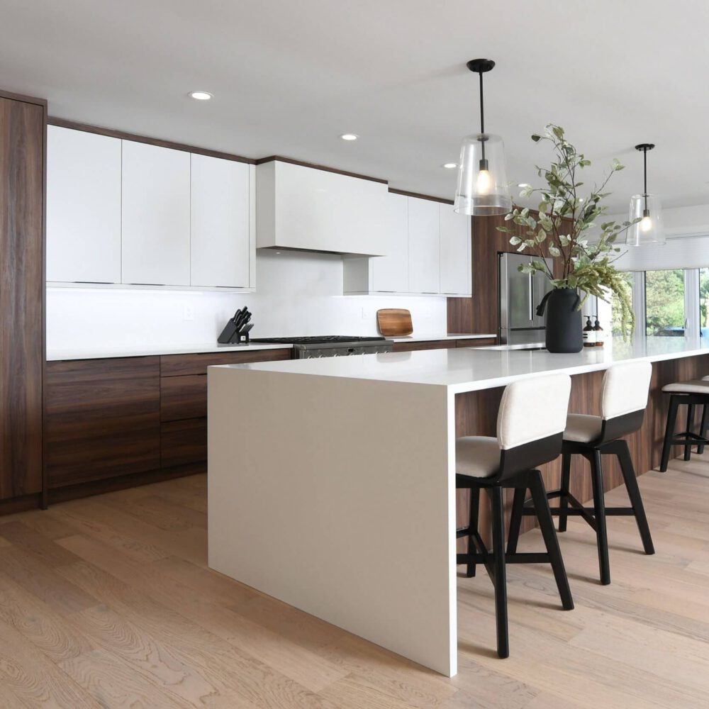 Casey’s Kitchens modern kitchen with white upper cabinets, dark wood lower panels, a large white island, and black-stem stools beneath pendant lights, featuring greenery in a black vase.