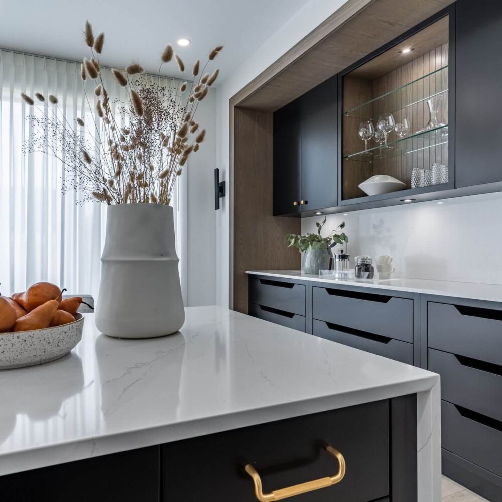 A modern kitchen shows dark cabinetry, a white quartz island, warm oak accents, and dried stems beside a bowl of fruit.