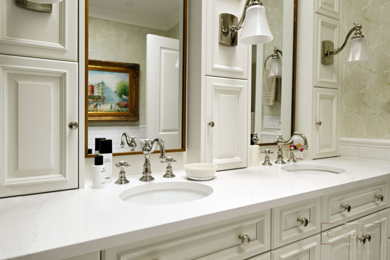 White bathroom countertop with two sinks, silver faucets, and a mirror above, featuring storage cabinets and decorative elements.